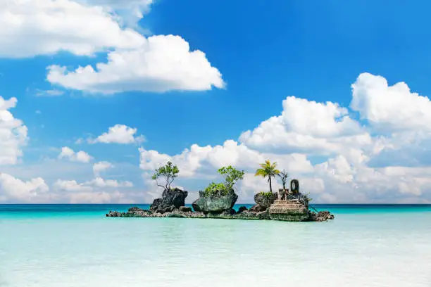 Willy's Rock, a famous rock formation situated on Boracay Island's White Beach. Positioned at the center of the frame, the rock stands as a prominent feature. The surrounding sea appears calm and transparent, exhibiting turquoise and dark blue tones towards the horizon. Small ripples gently disrupt the water surface. The sky above is a vibrant shade of blue, adorned with fluffy clouds. Notably, no individuals are visible in the photo, allowing the focus to remain on the natural beauty of the location.