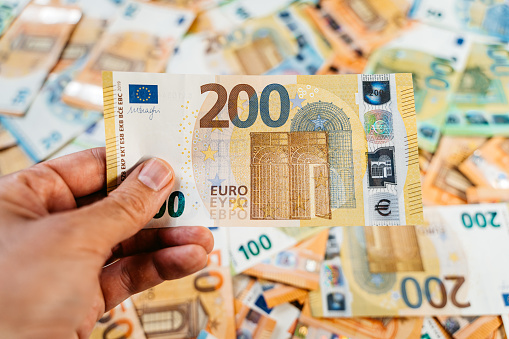 Male hand holding a single two hundred Euro above heap of assorted colorful EURO bills laid down on a flat surface.