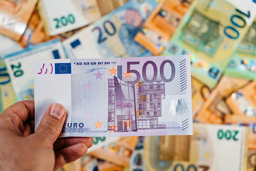 Male hand holding a single five hundred Euro above heap of assorted colorful EURO bills laid down on a flat surface.