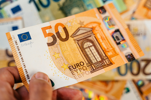 Male hand holding a single fifty Euro above heap of assorted colorful EURO bills laid down on a flat surface.