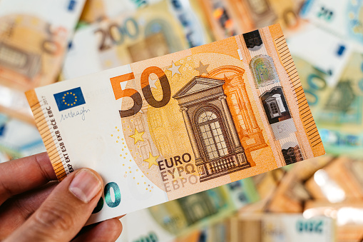 Male hand holding a single fifty Euro above heap of assorted colorful EURO bills laid down on a flat surface.