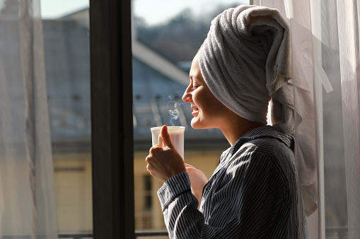 Beautiful young woman with bath towel on head holds a white cup of hot coffee or tea at home by the window. Smiling girl after shower is enjoying aroma drinks and have breakfast in the morning.
