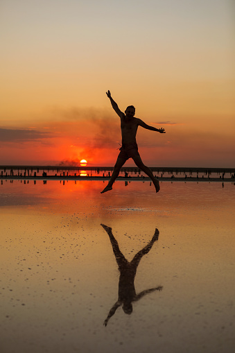 Summer tropic holiday vacation. Silhouette of young man by the sea at sunset having fun jumping enjoying freedom and life. Concept of travel wellbeing, happiness, success. Male reflection on water.