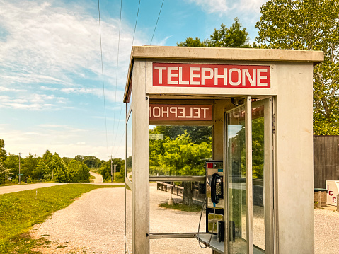 Vintage telephone booth situated on the side of a country road in rural Southern Illinois, USA. Gravel driveway, green leafy trees and a cloudy blue sky in the background of view.