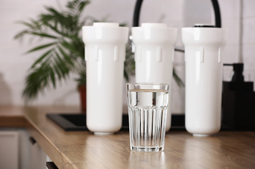 Glass of clean drinkable water and set of filter cartridges on wooden table top in a kitchen, houseplant.Installation of reverse osmosis water purification system. Concept household filtration system.