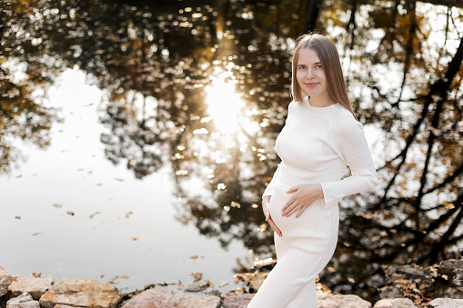 Mothers day. Beautiful young pregnant woman in white tight knit dress with little belly relaxing outdoors by the pond with reflection nature and sun in the park and looking at camera. Women day.