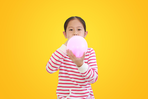 Pretty Asian little child girl blowing balloon isolated on yellow background with clipping path