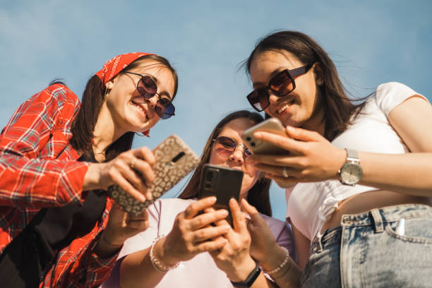 groupe de jeunes utilisant un appareil de téléphone portable debout ensemble à l’extérieur. les amis de la génération y accros aux applications de médias sociaux, aux paris ou aux jeux vidéo sur la plate-forme en ligne. - circle wireless technology application software group of people photos et images de collection