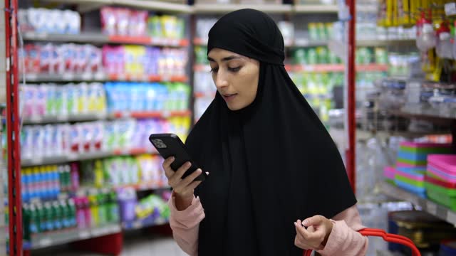 Portrait of Muslim woman in hijab with smartphone in hands in middle of supermarket. Woman in store chats with phone, consulting about purchases. Islamic woman with phone near store shelves with goods