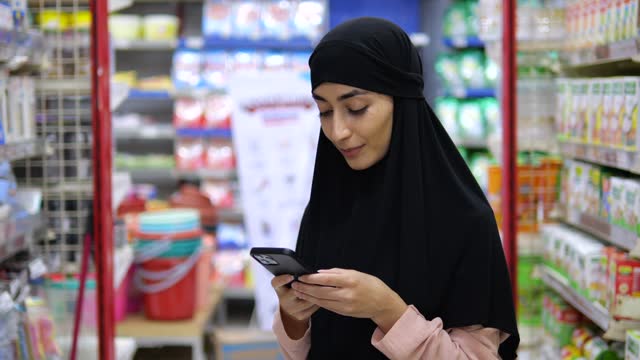 Beautiful woman in hijab in middle of the store shelves holds mobile phone in her hands answers message and smiles. Woman likes to talk on phone with her interlocutor, experiencing positive emotions.