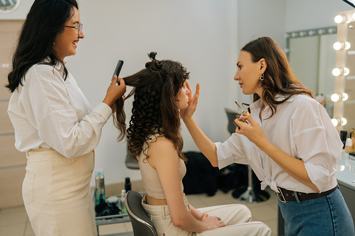 Side view of female makeup artist applying highlighter for face sculpting of pretty young woman in modern beauty studio. Smiling hairdresser brushing hair of lady during four-handed makeup and styling