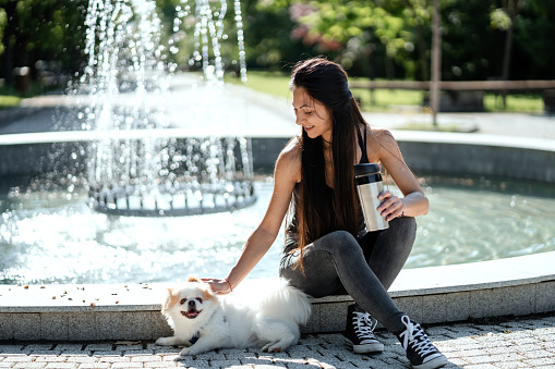 Young woman drinking coffee in front of fountain with her dog in the park