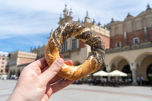 Hand holding obwarzanek krakowski pretzel on Kraków Main Market Square, with Cracow Cloth Hall Sukiennice in background. Krakowskie obwarzanki, traditional polish snack in Krakow, Poland.