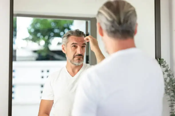 Handsome Caucasian grey-haired man with beard looks in the mirror healthy skin at home. Men's personal care. Middle aged man taking care of himself. Morning routine. Haircare and Lifestyle concept