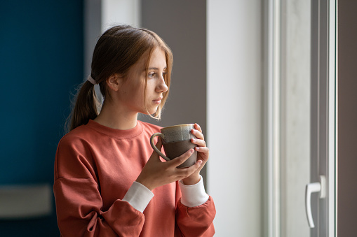 Thoughtful calm teen girl pondering teenage life standing with cup at home. Pensive schoolgirl looking at window, holding mug of hot drink, feeling loneliness without support friends. Difficult age.