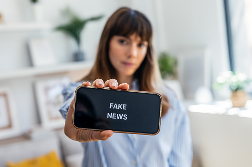 Shot of serious woman holding the mobile phone with the message fake news at home