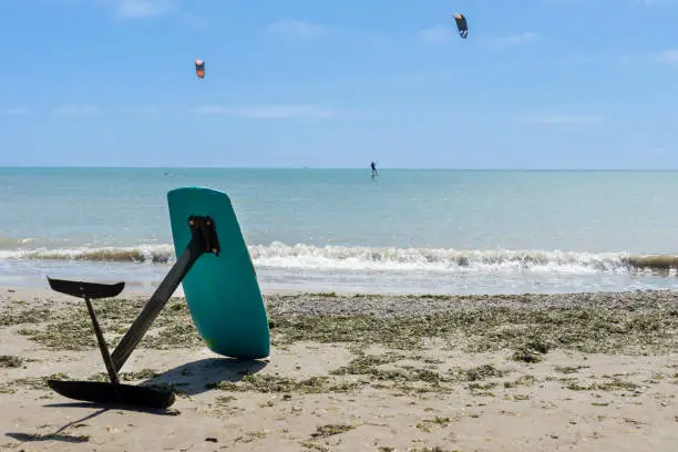 A kitesurf board resting alone on the sand of the beach with two kitesurfers surfing in the background. Water sport concept