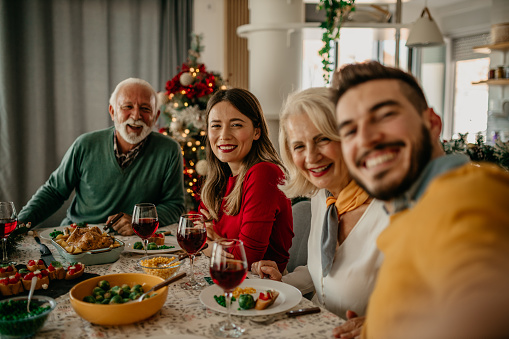 Smiling faces surround a table adorned with delicious food, taking a selfie as a family celebrates Christmas lunch in merry spirits.