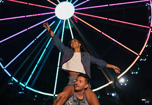Happy woman sitting on the shoulders of her boyfriend and having fun. Young couple against ferris wheel during the festival.