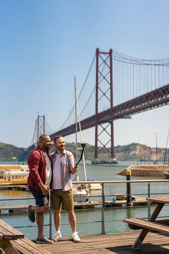 Gay couple taking a selfie in front of the 25 de Abril Bridge in Lisbon, Portugal