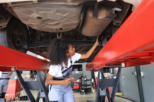 A young female mechanic, in uniform, holds a tablet in her hands and inspects the car. Car on the lift. Rear and bottom view, Female car mechanic working under a car on a lift,  Low angle shot of a woman auto technician examining lifted automobile. Profession, occupation concept