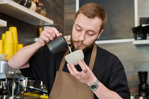 Male barista preparing coffee in coffee shop, bearded young man worker in an apron standing near coffee machine. Small business, service, coffee shop, staff, work concept