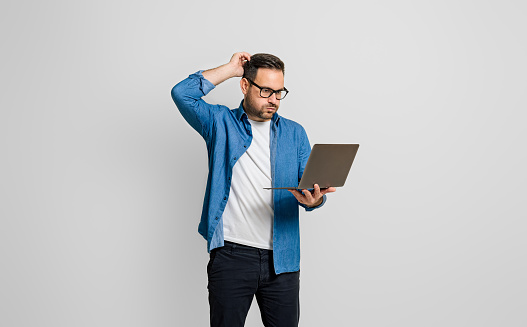 Confused disappointed businessman with hand in hair reading e-mail over laptop on gray background