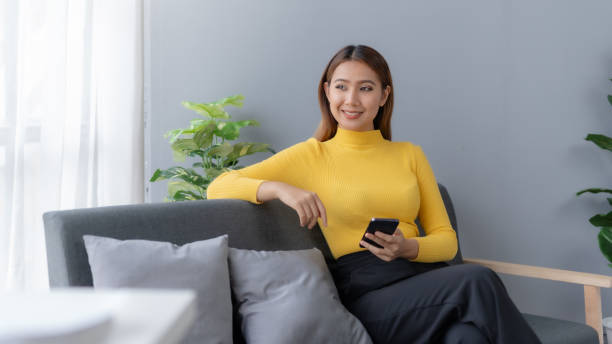 istock Modern Asian Woman Using Smartphone at Home. She sits comfortably on a sofa at home, browsing on her smartphone. 1505975860