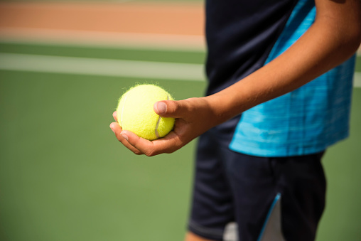 Close-up on boy's hand holding tennis ball on court outdoors. Kid is 10 year’s old, and wearing a deep blue t-shirt and shorts. No face. Horizontal close-up outdoors shot with copy space. This was taken in Florida, USA.