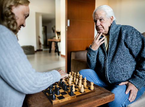 Senior man and his wife sitting together and playing a game of chess at a table at home