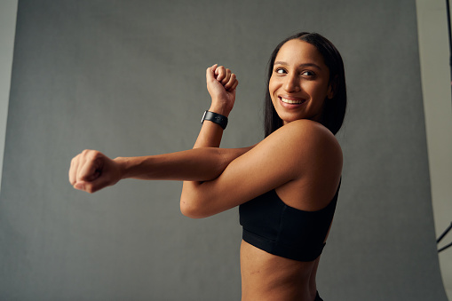 Young biracial woman wearing sports bra looking away and smiling while doing cross arm stretch in studio