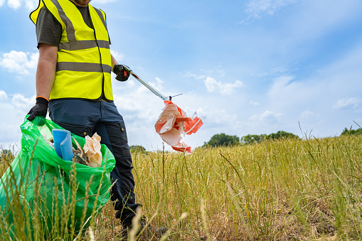 Young man picking up litter in the nature with a reflective vest, trash bag and a litter picker.