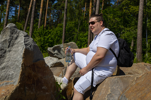 In summer, a handsome man in white clothes sits on a stone with a bottle of water and takes a break from the summer heat. The traveler regains strength after a long journey..