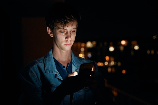 Focused young caucasian man in casual clothing texting on mobile phone outside apartment at night