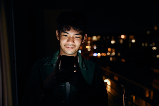 Young multiracial man wearing shirt text messaging with mobile phone on balcony of apartment at night