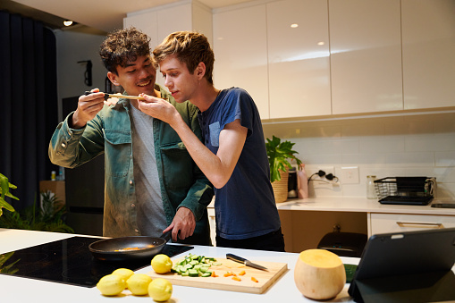 Young gay couple in casual clothing smiling while tasting dinner by digital tablet in kitchen at home