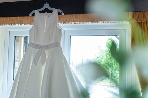 A white wedding dress hangs from a clothing rack in front of a bright window, illuminated by warm, natural light