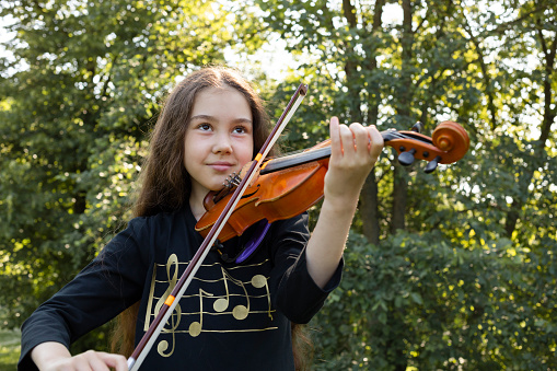 Little Smiling Beautiful Girl Plays On Violin, Musical String Instrument, Green Park And Sun On Background. Little Asian Caucasian Child, Talented Artist, Horizontal Plane. Lifestyle.