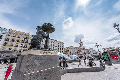 Madrid, Spain - June 20, 2023: Tourists taking photos of the sculpture The Bear and the Strawberry Tree, symbol of the city of Madrid, in the Plaza de la Puerta del Sol. This a pedestrian public square in Madrid, one of the best known and busiest places in the city. This point marks the zero kilometers of the radial network of Spanish roads. The Puerta del Sol originated as one of the gates in the city wall that surrounded Madrid in the 15th century.