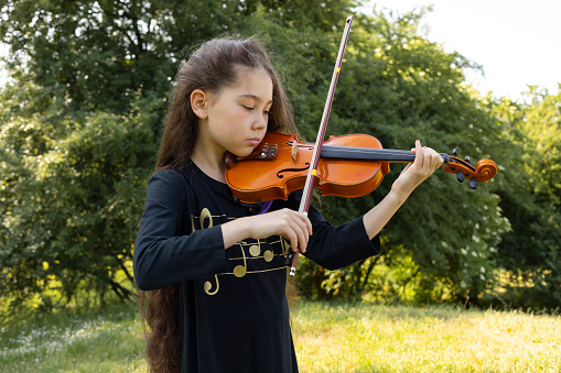 Asian Caucasian Child Plays On Violin, Musical String Instrument, Green Park On Background. Little Adorable Child, Talented Artist, Lifestyle. Horizontal Plane. . High quality photo