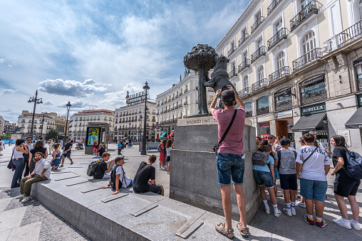 Madrid, Spain - June 20, 2023: Tourists taking photos of the sculpture The Bear and the Strawberry Tree, symbol of the city of Madrid, in the Plaza de la Puerta del Sol. This a pedestrian public square in Madrid, one of the best known and busiest places in the city. This point marks the zero kilometers of the radial network of Spanish roads. The Puerta del Sol originated as one of the gates in the city wall that surrounded Madrid in the 15th century.