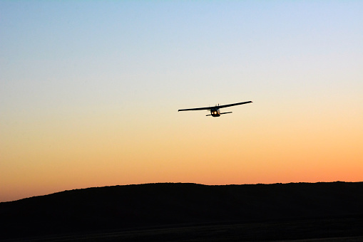 A lone small single-engine plane flies in the clear evening sky. Only the outline of the plane and the ground below are visible. Front view
