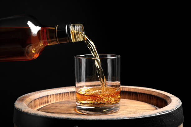 Pouring whiskey from bottle into glass on wooden barrel against black background, closeup stock photo