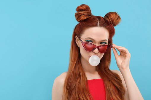 Beautiful woman in sunglasses blowing bubble gum on light blue background. Space for text