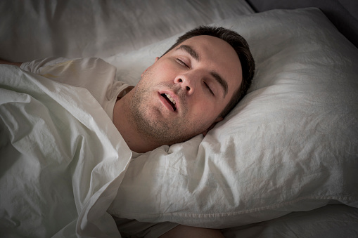 View Of Tired Young Man Snoring While Deep Sleeping In Bed. The problem of snoring during sleep. A young cute guy sleeps on a white bed at night