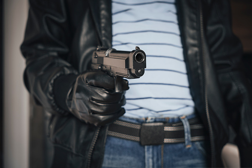 Give me your money. robber aiming with pistol the victim. Hooded armed man thief killer in leather jacket holds gun in gloved hand. Weapon, crime.