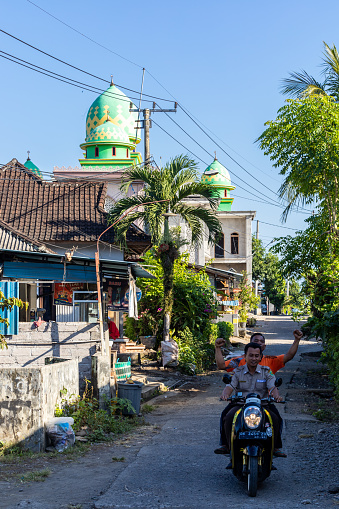Gilimanuk, Bali, Indonesia, May 27, 2023  A policeman and passenger in a back alley riding a motorcycle with a mosque in the background.