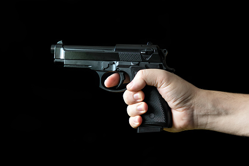 Hand holding a gun, on black background. The concept of the crime of banditry. A dangerous shooter and a black pistol on a dark background. The hired killer is preparing to shoot.