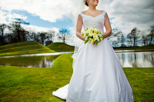 Happy blond woman wearing weeding dress and holding bouquet. Studio shot against grey background.