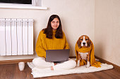 A smiling girl in warm clothes is sitting on the floor in the house with her dog. A teenager is working on a laptop.